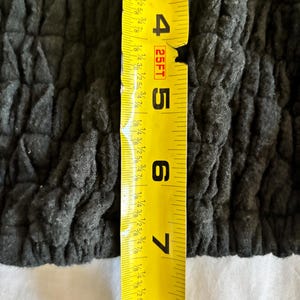May include: Close-up of a dark gray, textured fabric. A yellow measuring tape is placed vertically, showing measurements from 3 to 8 inches. The fabric appears to be a garment or textile.