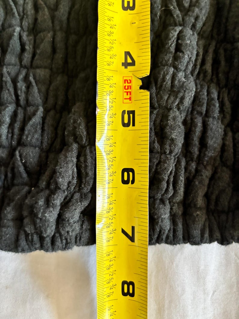 May include: Close-up of a dark gray, textured fabric. A yellow measuring tape is placed vertically, showing measurements from 3 to 8 inches. The fabric appears to be a garment or textile.