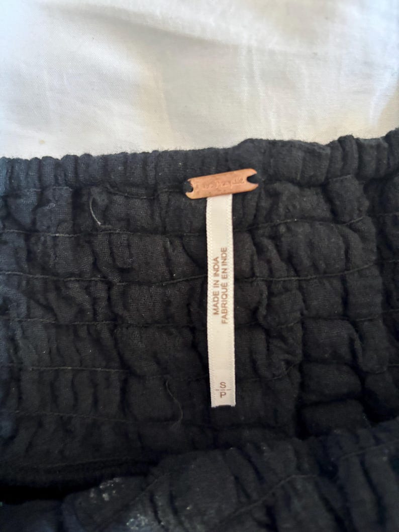 May include: Close-up of a black garment with a textured, gathered design. A small, brown leather tag with the word "Free People" is attached. A white tag with text that reads "MADE IN INDIA FABRIQUE EN INDE" is also visible.