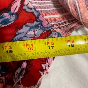 May include: Close-up of a fabric with a floral pattern in red, pink, and blue. The fabric is measured with a yellow tape measure, showing a length of approximately 15 to 18 inches.