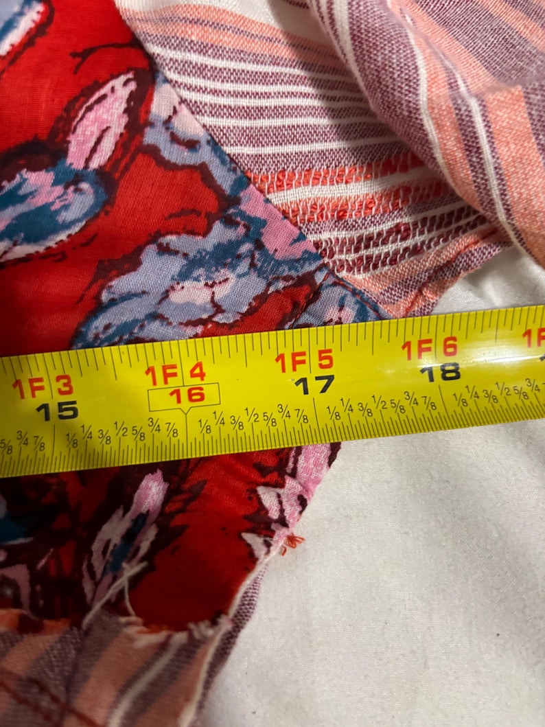 May include: Close-up of a fabric with a floral pattern in red, pink, and blue. The fabric is measured with a yellow tape measure, showing a length of approximately 15 to 18 inches.