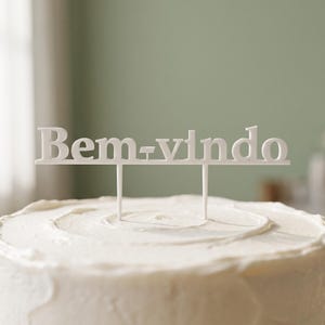 Puede incluir: Un adorno para pastel de color blanco con el texto "Bem-vindo" en una fuente con serifa, sostenido por dos varillas verticales delgadas. El adorno está colocado en posición vertical sobre un pastel redondo cubierto de glaseado blanco liso.