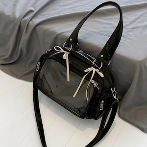 May include: Black patent leather handbag with a clear front panel. The bag features a top handle, a detachable shoulder strap, and white ribbon accents. Silver-toned hardware and zippers complete the design.