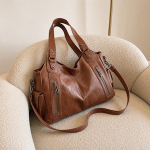 May include: A brown handbag with a smooth texture, featuring two top handles and a detachable shoulder strap. The bag includes two vertical zippered pockets on the front, side pockets, and silver-toned metal hardware accents on the strap attachments.