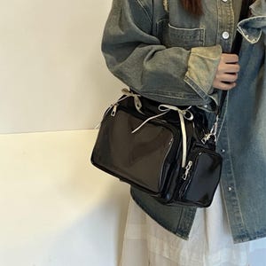 May include: A black patent leather crossbody bag with multiple compartments and a small, attached pouch. The bag features silver-toned hardware, a long adjustable strap, and a white ribbon detail. The bag is designed for carrying essentials.