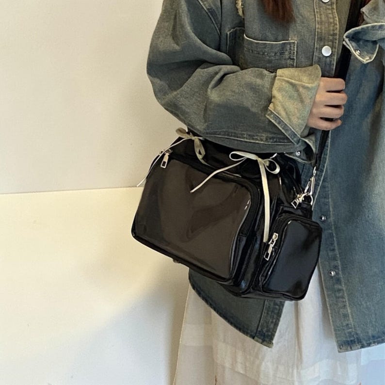 May include: A black patent leather crossbody bag with multiple compartments and a small, attached pouch. The bag features silver-toned hardware, a long adjustable strap, and a white ribbon detail. The bag is designed for carrying essentials.