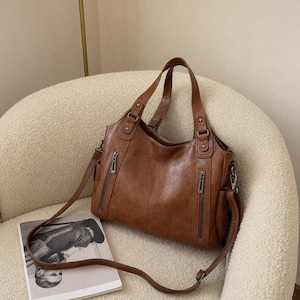 May include: A brown leather handbag featuring two vertical zippered pockets on the front, dual top handles, and a detachable shoulder strap. The bag has a structured design with visible stitching, metallic hardware, and side pockets for storage.