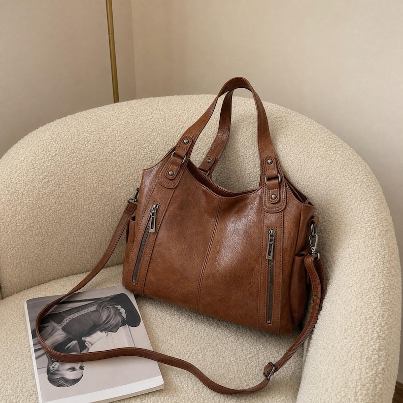 May include: A brown leather handbag featuring two vertical zippered pockets on the front, dual top handles, and a detachable shoulder strap. The bag has a structured design with visible stitching, metallic hardware, and side pockets for storage.