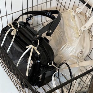 May include: Two handbags in a metal basket. One is black with white ribbon accents, a top handle, and a shoulder strap. The other is white with a clear front panel and a shoulder strap. Both have silver hardware.