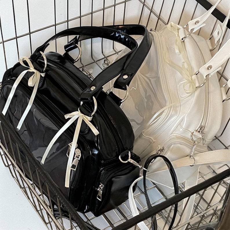 May include: Two handbags in a metal basket. One is black with white ribbon accents, a top handle, and a shoulder strap. The other is white with a clear front panel and a shoulder strap. Both have silver hardware.