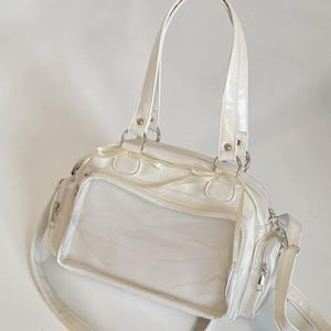 May include: White handbag with a clear front panel, featuring a top handle and a detachable shoulder strap. The bag has silver-toned hardware, a zippered side pocket, and decorative ribbon accents.