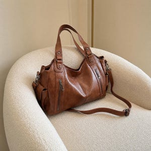 May include: A medium-sized brown handbag with a smooth finish, featuring two top handles and a detachable shoulder strap. The bag includes two vertical zippered pockets on the front, side pockets, and silver-toned metal hardware accents.