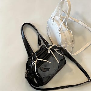 May include: Two handbags, one black and one white, are displayed against a neutral background. The black bag has a glossy finish, a clear front panel, and white ribbon accents. The white bag has a rounded shape and a top handle. Both bags have shoulder straps.