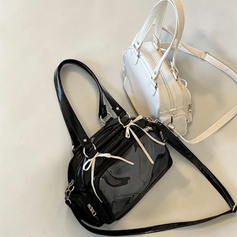 May include: Two handbags, one black and one white, are displayed against a neutral background. The black bag has a glossy finish, a clear front panel, and white ribbon accents. The white bag has a rounded shape and a top handle. Both bags have shoulder straps.