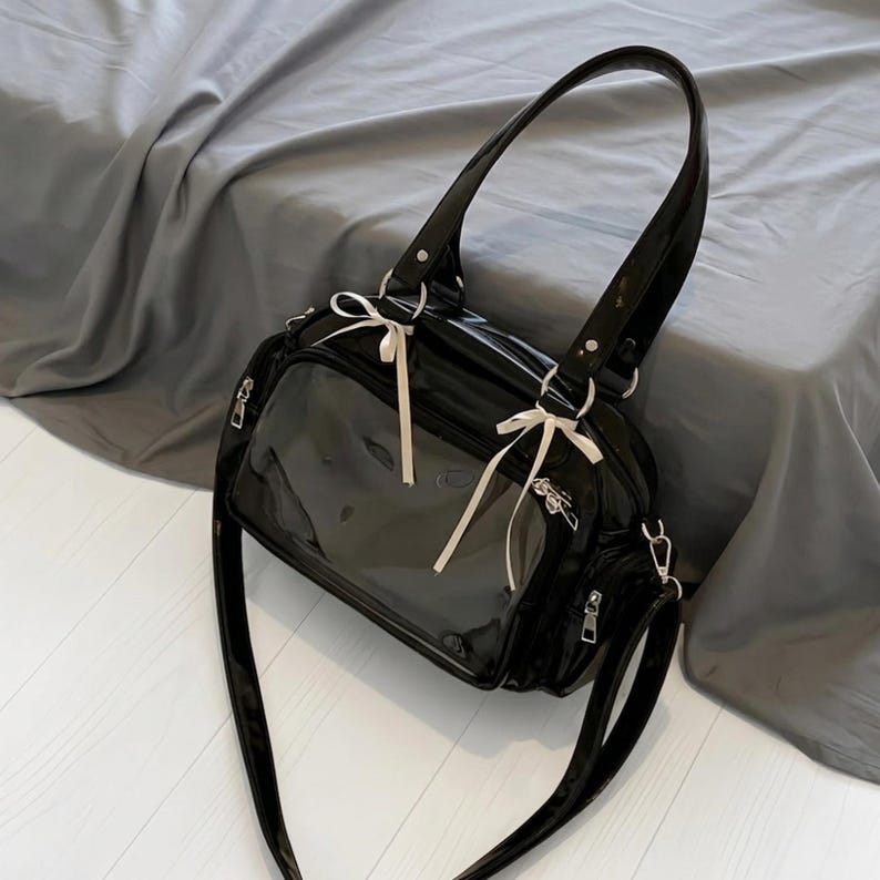 May include: Black patent leather handbag with a clear front panel. The bag features a top handle, a detachable shoulder strap, and white ribbon accents. Silver-toned hardware and zippers complete the design.