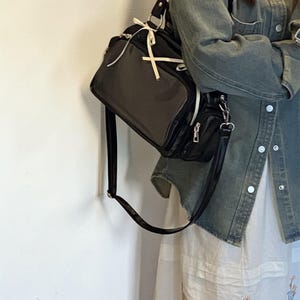 May include: Black shoulder bag with a rectangular shape and a smaller attached pouch. The bag has a long strap and white accents. The bag is hanging against a white wall. The bag is made of a shiny material.