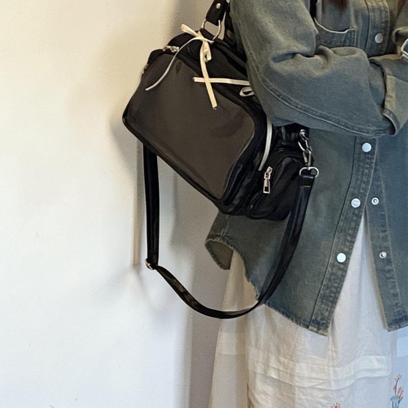 May include: Black shoulder bag with a rectangular shape and a smaller attached pouch. The bag has a long strap and white accents. The bag is hanging against a white wall. The bag is made of a shiny material.