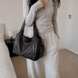 May include: A dark brown leather handbag featuring two top handles, two vertical zippered pockets on the front, and a side pocket with a flap closure. The bag has a structured base and silver-toned hardware accents on the handle attachments and zippers.