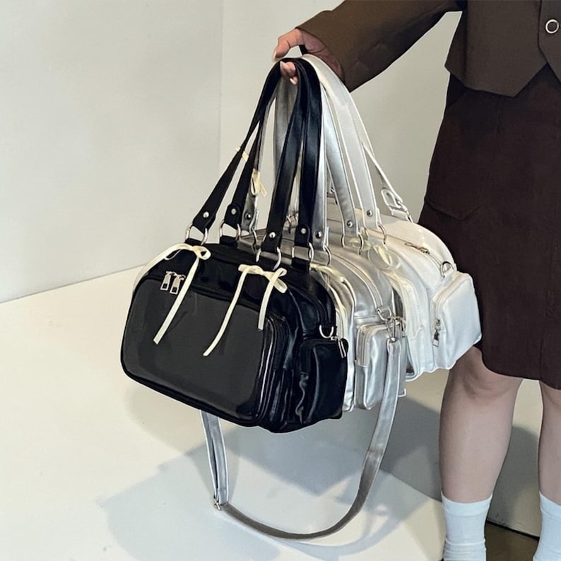 May include: A collection of three shoulder bags in black, silver, and white. Each bag features a rectangular shape, multiple compartments, and a long, adjustable strap. The bags have silver hardware and white accents.
