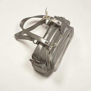 May include: A silver handbag with a glossy finish. The bag features a top handle, a zippered main compartment, and an additional front pocket. White ribbons are tied to the handle and zipper pulls. The bag is designed for carrying essentials.