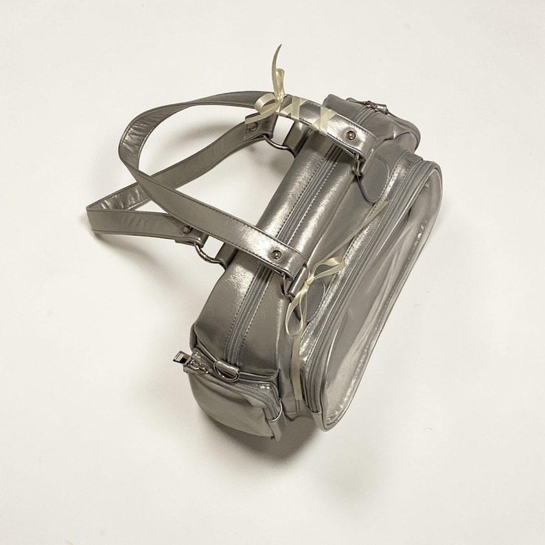 May include: A silver handbag with a glossy finish. The bag features a top handle, a zippered main compartment, and an additional front pocket. White ribbons are tied to the handle and zipper pulls. The bag is designed for carrying essentials.