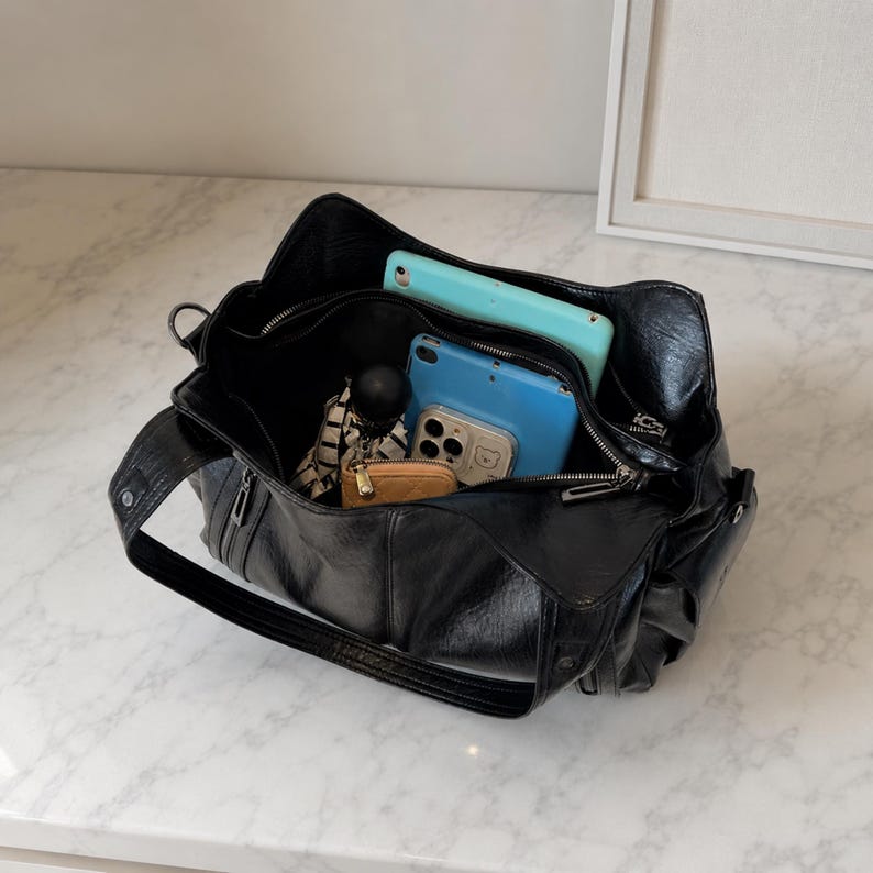 May include: A black faux leather shoulder bag with a wide strap and zippered closure. The interior contains a light blue tablet, a smaller blue electronic device, a beige wallet, and a patterned fabric accessory, all visible within the open bag.