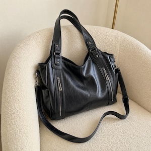 May include: A black shoulder bag with a textured finish, featuring two top handles and a detachable crossbody strap. The front includes two vertical zippered pockets with silver-toned hardware, side slip pockets, and reinforced handle attachments.