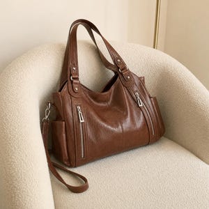 May include: A brown leather handbag featuring two vertical zippered pockets on the front, two top handles with silver-toned metal hardware, and a detachable shoulder strap. The bag includes side pockets and a smooth, textured finish throughout.