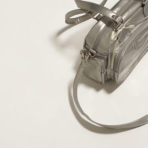 May include: Silver handbag with a shoulder strap and top handles. The bag has a rectangular shape with multiple compartments and a zipper closure. The material appears to be shiny and reflective, with a metallic finish. The bag is set against a white background.