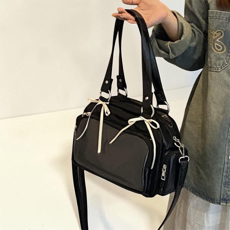 May include: Black handbag with a glossy finish, featuring a rectangular shape and dual handles. The bag has a transparent panel and is accented with white bows. It includes a shoulder strap and a small side pocket with a zipper.