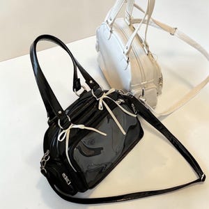 May include: Two handbags. The front bag is black with a glossy finish, a clear panel, and white ribbon accents. The second bag is white with a similar shape and design. Both bags have handles and shoulder straps, and are on a white surface.