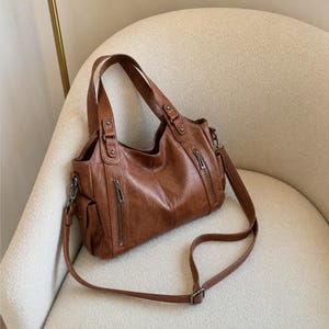 May include: A brown handbag with a textured finish, featuring two top handles and a detachable shoulder strap. The bag includes two vertical zippered pockets on the front, side pockets, and silver-toned metal hardware attachments for the straps.