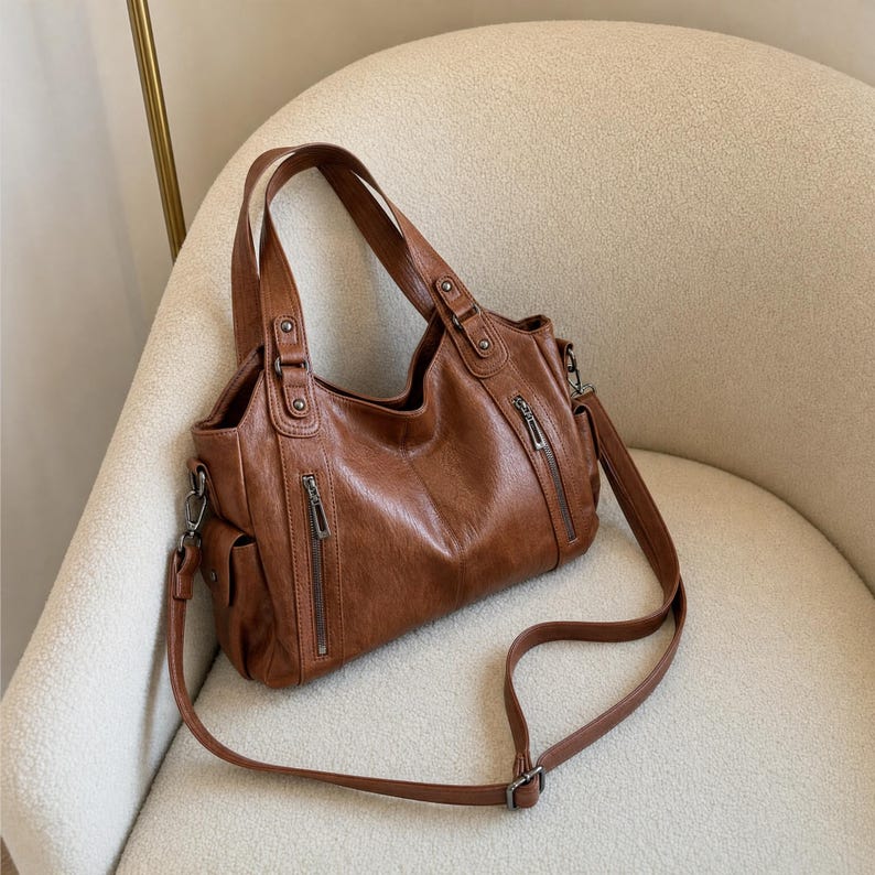 May include: A brown handbag with a textured finish, featuring two top handles and a detachable shoulder strap. The bag includes two vertical zippered pockets on the front, side pockets, and silver-toned metal hardware attachments for the straps.