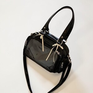 May include: Black handbag with a clear front panel, featuring a glossy finish and a shoulder strap. The bag has a top handle and is adorned with cream-colored bows. Silver zippers and hardware add detail to the design.