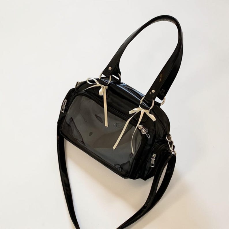 May include: Black handbag with a clear front panel, featuring a glossy finish and a shoulder strap. The bag has a top handle and is adorned with cream-colored bows. Silver zippers and hardware add detail to the design.