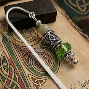 May include: A silver bookmark with a decorative bead design. The beads include a green gemstone, a silver Celtic knot charm, and a light green bead. The bookmark is resting on a book with Celtic knot designs.