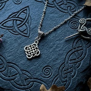 May include: A silver-toned metal pendant featuring a square-shaped Celtic knot design suspended from a delicate matching chain. The pendant is positioned near a silver-toned metal brooch with a spiral pattern on a dark textured surface.