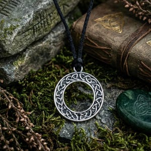 A silver-colored pendant with a Celtic knot design hangs from a black cord. The circular pendant is intricately detailed. The background features moss, rocks, and a leather-bound book, suggesting a historical or fantasy theme.