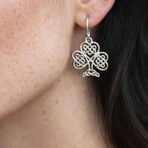 May include: A silver-toned earring featuring a three-lobed shamrock design with intricate, interwoven Celtic knotwork patterns. The metal charm hangs from a simple silver-toned hook, displaying a polished finish and symmetrical, open-work detailing.