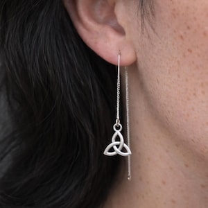 Pode incluir: Um brinco de fio prateado com uma corrente delicada que passa pelo lóbulo da orelha e termina num pequeno pendente de triquetra de três lóbulos. O metal tem um acabamento polido e o design pende verticalmente a partir do furo.