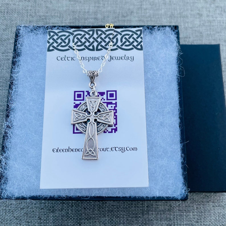 A silver Celtic cross pendant necklace with intricate knotwork designs. The necklace is displayed in a black gift box with white tissue paper. The box has a white card with the text "Celtic Inspired Jewelry" and a QR code.