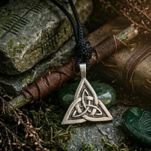 May include: A silver-toned metal pendant featuring an intricate triquetra knot design, suspended from a black braided cord. The pendant has a triangular shape with interlaced lines and a polished finish, hanging from a knotted cord loop.