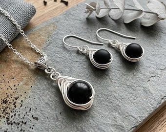 May include: A jewelry set consisting of a necklace and a pair of drop earrings. Each piece features a smooth, round black bead encased in silver-toned wire wrapping. The necklace pendant includes a triangular bail with a Celtic knot design.
