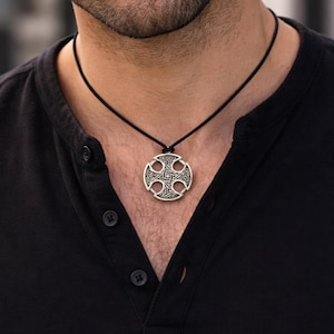 May include: A silver-toned circular pendant featuring an intricate Celtic knotwork cross design, suspended from a simple black cord necklace. The pendant has a metallic finish with dark recessed areas that highlight the detailed engraved patterns.