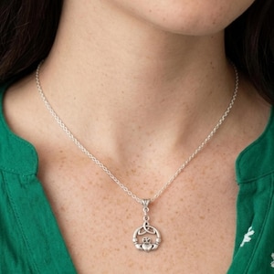 Puede incluir: Un collar con colgante Claddagh de tono plateado que presenta un corazón sostenido por dos manos y coronado, suspendido de una delicada cadena plateada. El diseño del colgante incorpora elementos de nudos celtas.