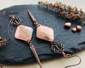 May include: A pair of copper earrings featuring a central diamond-shaped metal plate, a circular wire-wrapped knot design, and a long, pointed metal spike. The earrings are suspended from curved ear wires and have a polished, metallic finish.