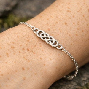 May include: A silver-toned bracelet featuring a central infinity symbol charm connected to a delicate chain. The metal charm displays a polished finish with a continuous looped design, secured by a simple link chain that wraps around the wrist.