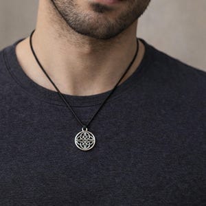 May include: A silver-toned circular pendant featuring an intricate Celtic triquetra knot design, suspended from a simple black cord necklace. The pendant has a polished metallic finish and rests against a dark charcoal gray crew neck t-shirt.