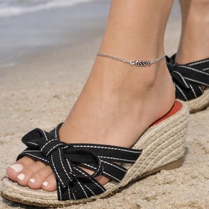 May include: A silver chain anklet featuring a central decorative metal link design, paired with black wedge sandals that have a jute-wrapped heel and a prominent fabric bow detail on the top strap with white contrast stitching throughout.