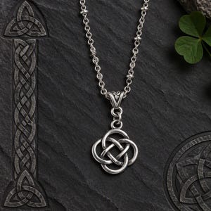 May include: A silver necklace featuring a pendant with an intricate four-fold Celtic knot design. The pendant hangs from a delicate silver chain with small spherical accents. The metal has a polished finish with darkened recessed areas.